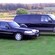 A town car parked next to an SUV on a grassy lawn near a road against a backdrop of grass, trees and a small lake